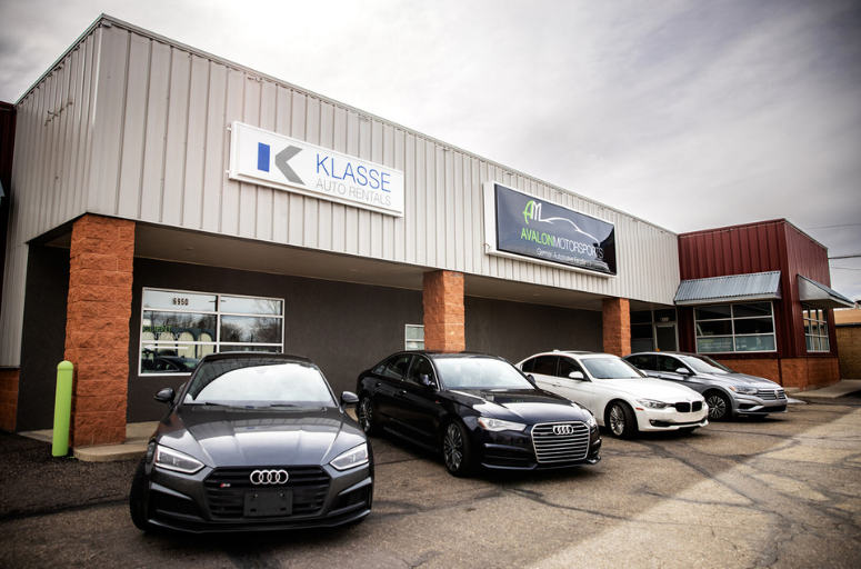 The exterior view of Avalon Motorsports in Denver, featuring premium vehicles such as Audis and BMWs parked in front of the building. Visible signage includes "Avalon Motorsports" and "Klasse Auto Rentals" on a clear day.