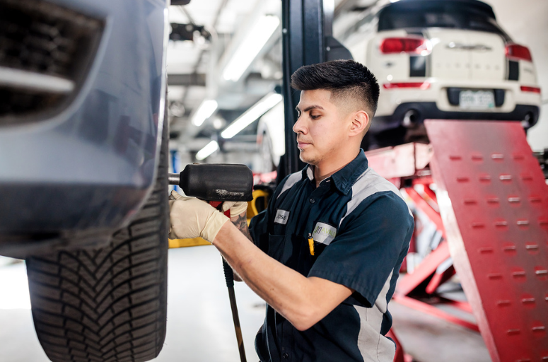 A skilled auto technician using a pneumatic tool to secure a vehicle's tire in a professional repair shop. The environment features modern equipment, a lift system, and vehicles in the background.